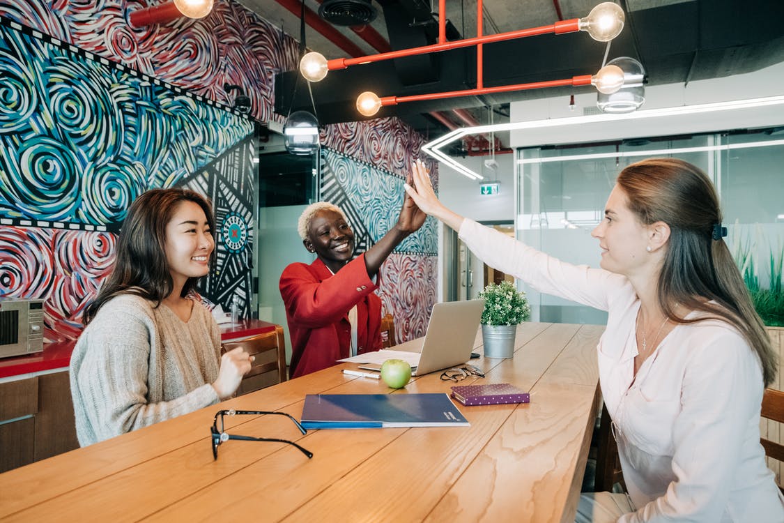 Group of positive young multiethnic female colleagues smiling and giving high five while sitting at wooden table with documents and laptop in creative workspace