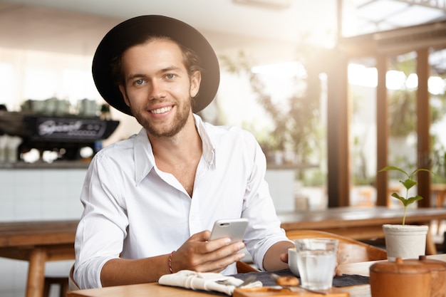 Free photo attractive happy young bearded man in trendy hat texting messages via social networks and browsing internet, using free wifi on his electronic device during coffee break at restaurant