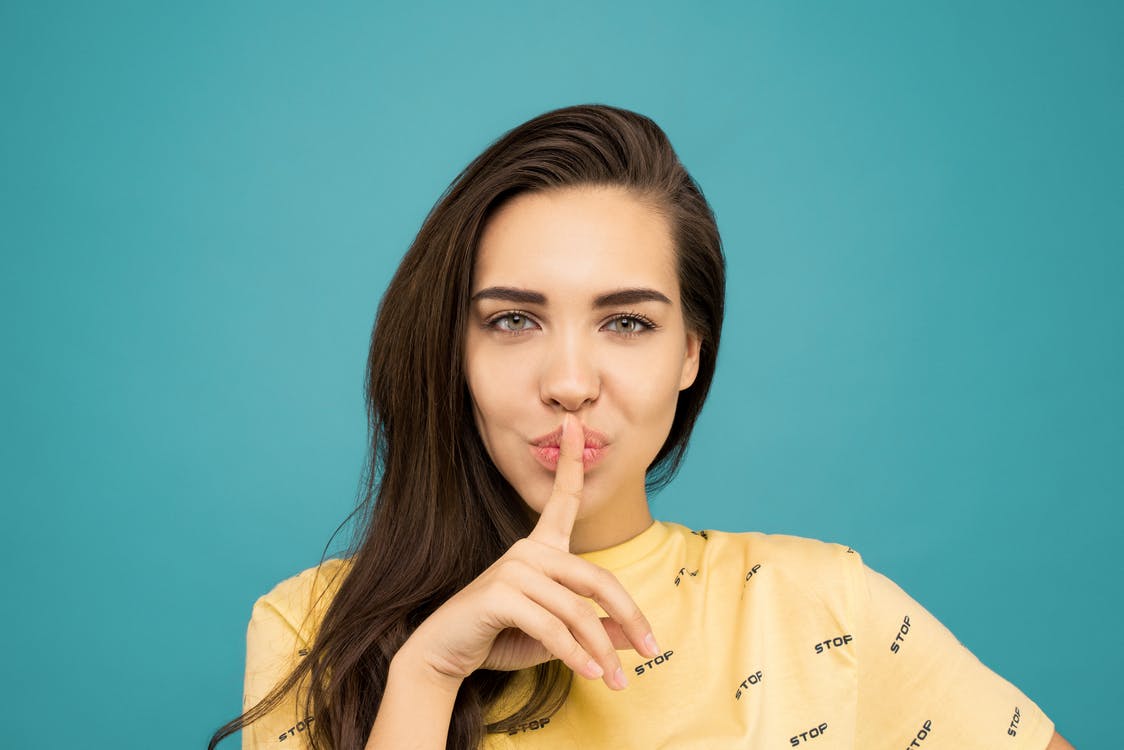 Portrait Photo of Woman in Yellow T-shirt Doing the Shh Sign While Standing In Front of Blue Background